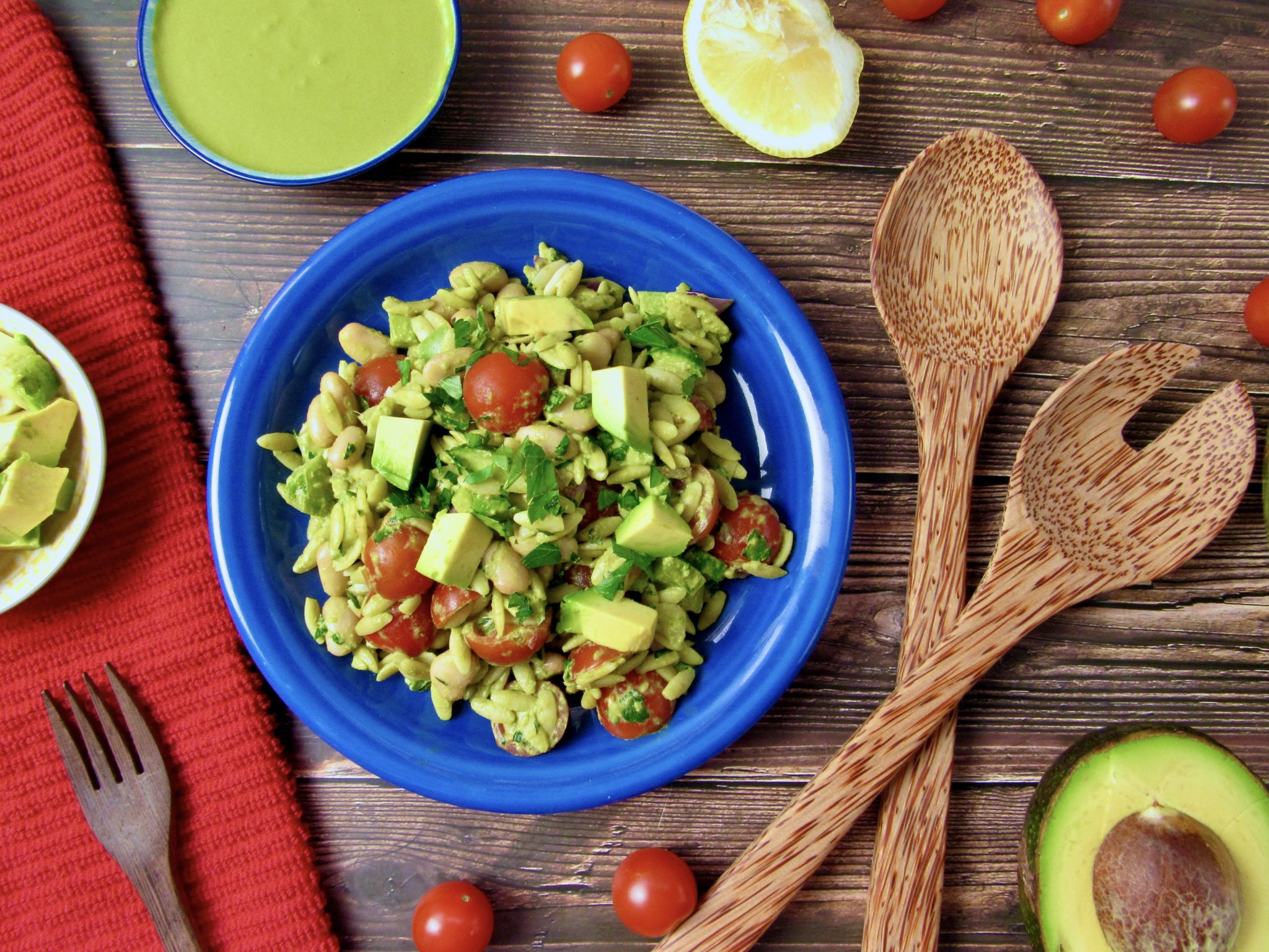 A vibrant pasta salad featuring diced avocado, cherry tomatoes, and fresh greens is served on a blue plate. Surrounding the plate are wooden utensils, a halved avocado, a lemon slice, a red cloth, and a small bowl of green dressing.