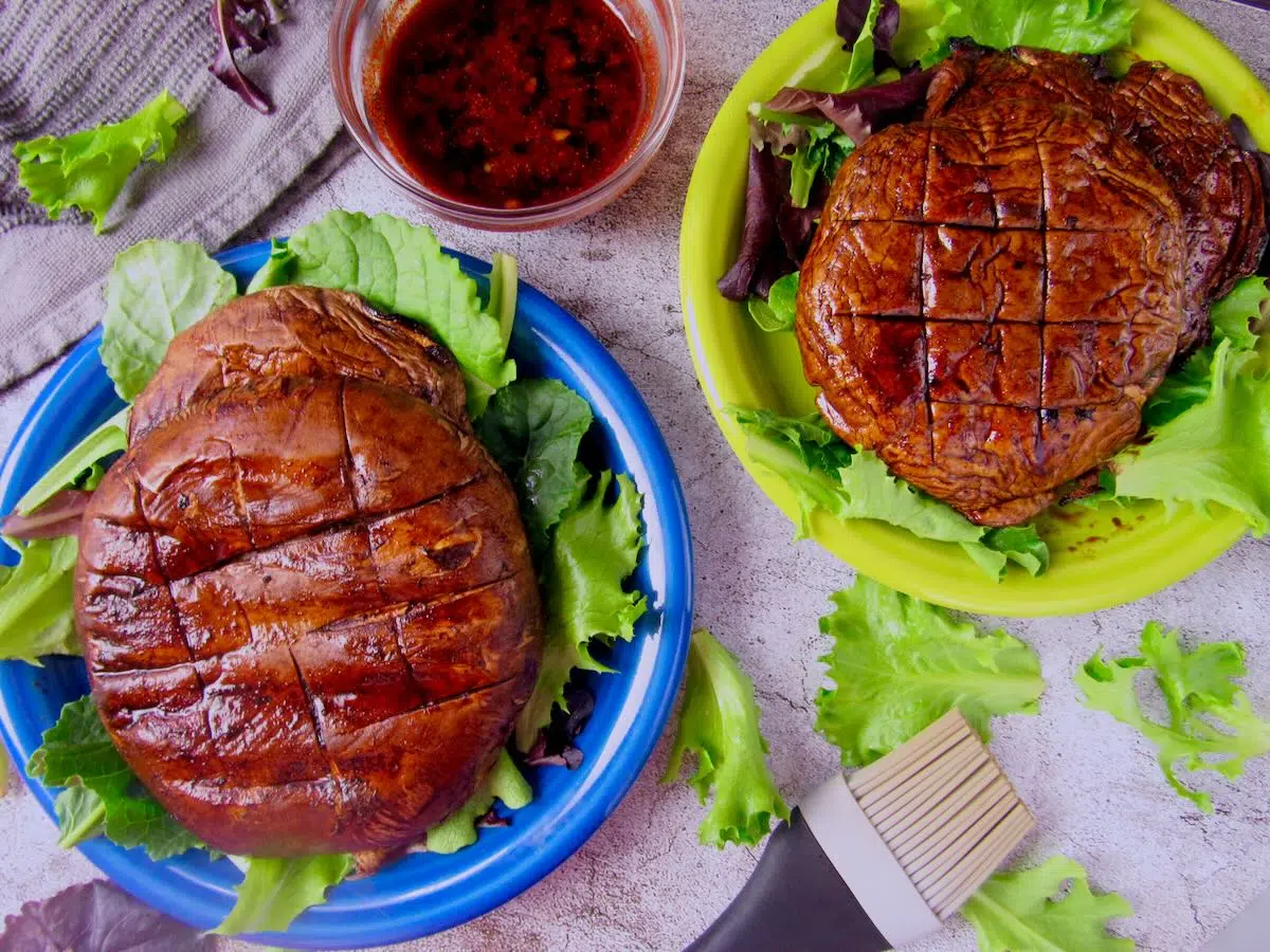 Two grilled portobello mushrooms on beds of leafy greens, accompanied by a small bowl of sauce and a brush, set on a light surface.