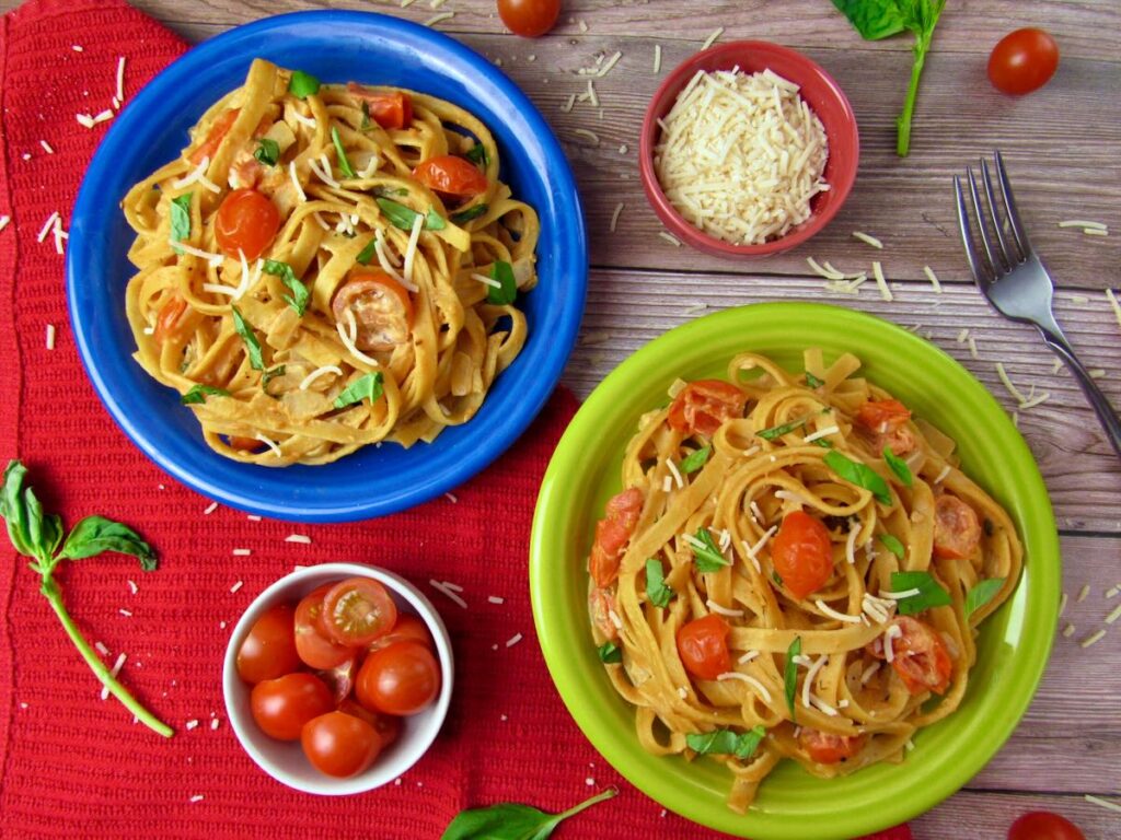 Two plates of creamy pasta topped with cherry tomatoes, shredded cheese, and herbs, are placed on a wooden table. One plate is blue, and the other is green. Surrounding the plates are small bowls of cherry tomatoes and shredded cheese, along with a fork and basil leaves.