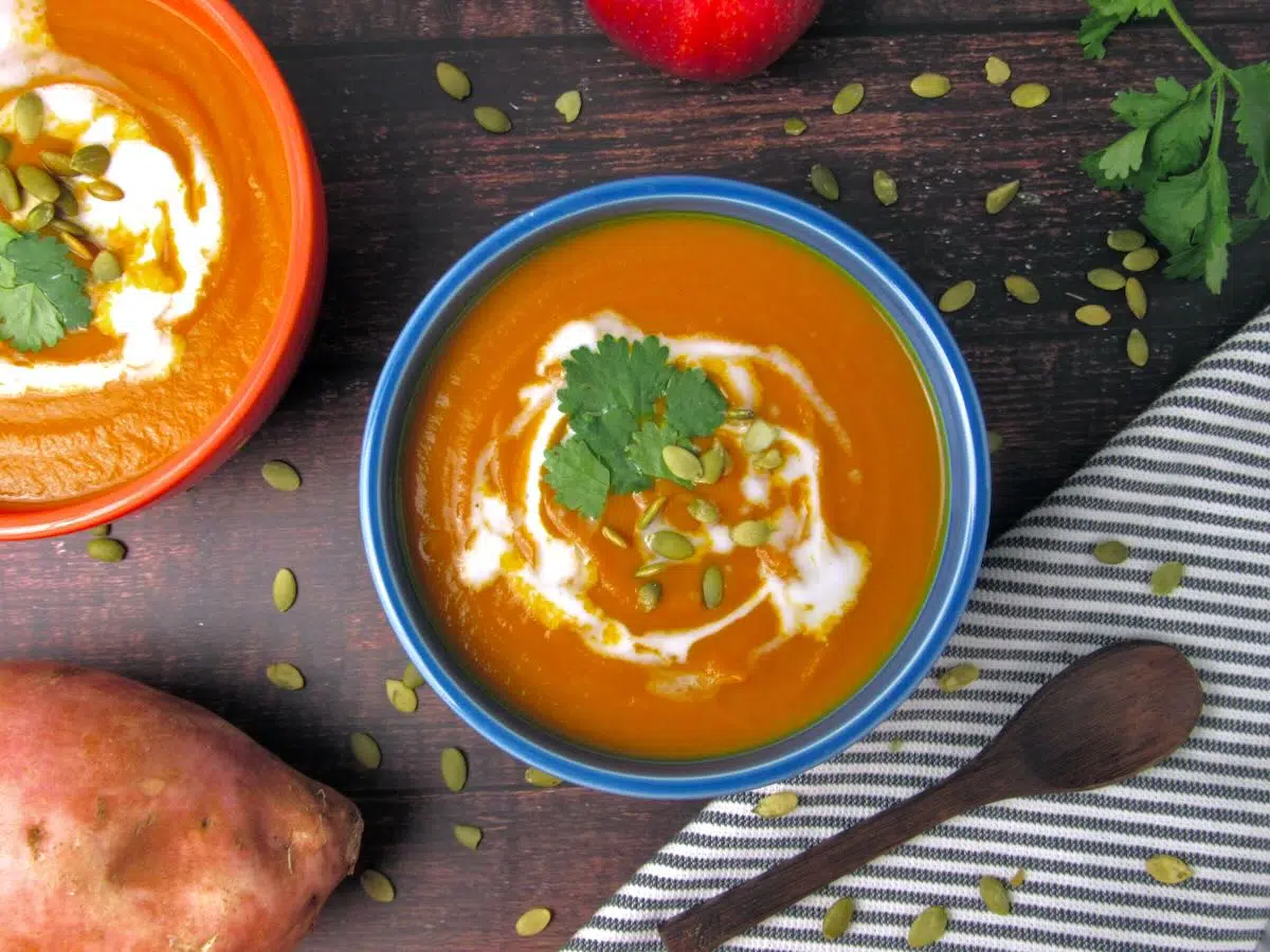 A bowl of creamy orange soup garnished with pumpkin seeds and cilantro, alongside another bowl, a red apple, a sweet potato, and a striped napkin on a dark wooden table.