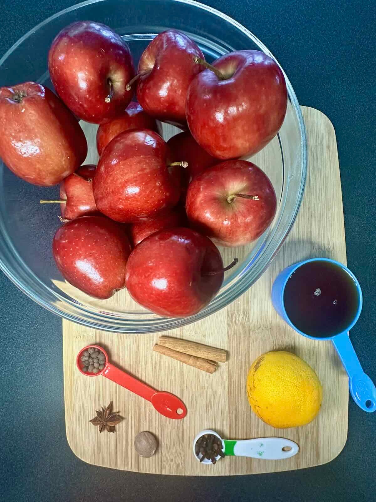 Ingredients for Homemade Spiced Apple Cider including fresh red apples, orange, maple syrup, cinnamon sticks, and whole spices on a wooden board.