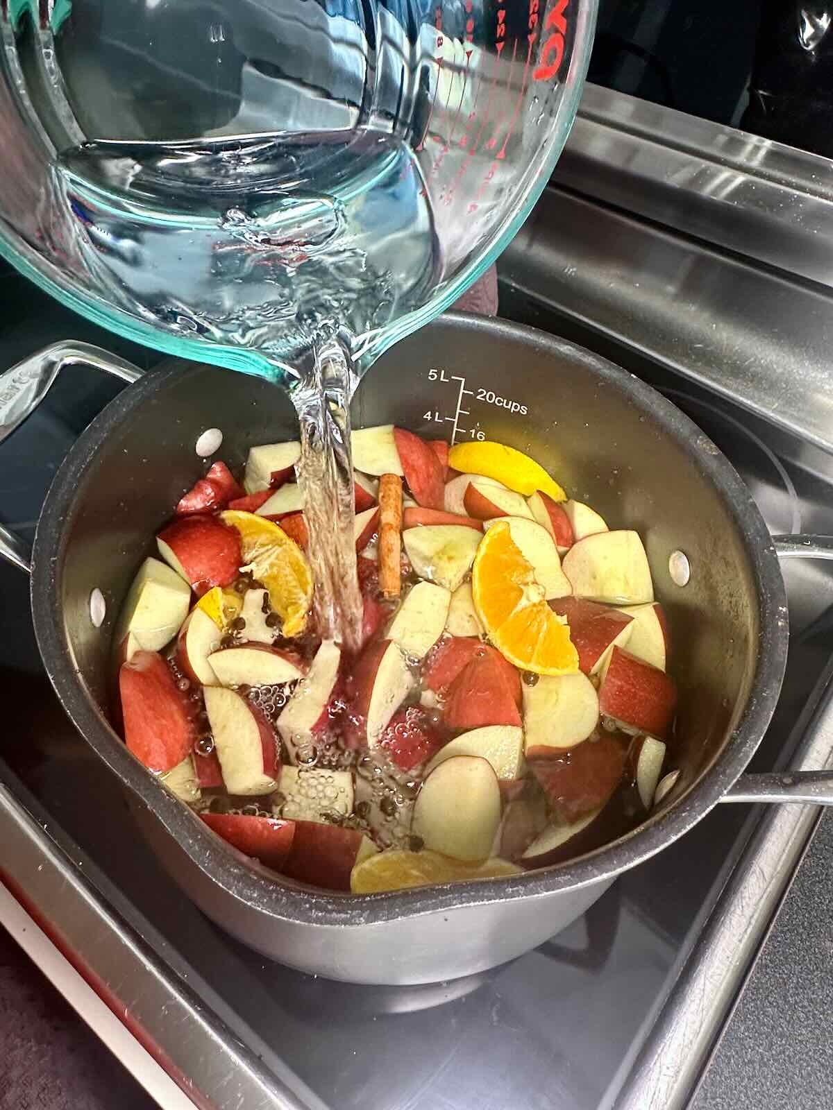Pouring water into a large pot filled with chopped apples, orange slices, cinnamon sticks, and spices to make Homemade Apple Cider.