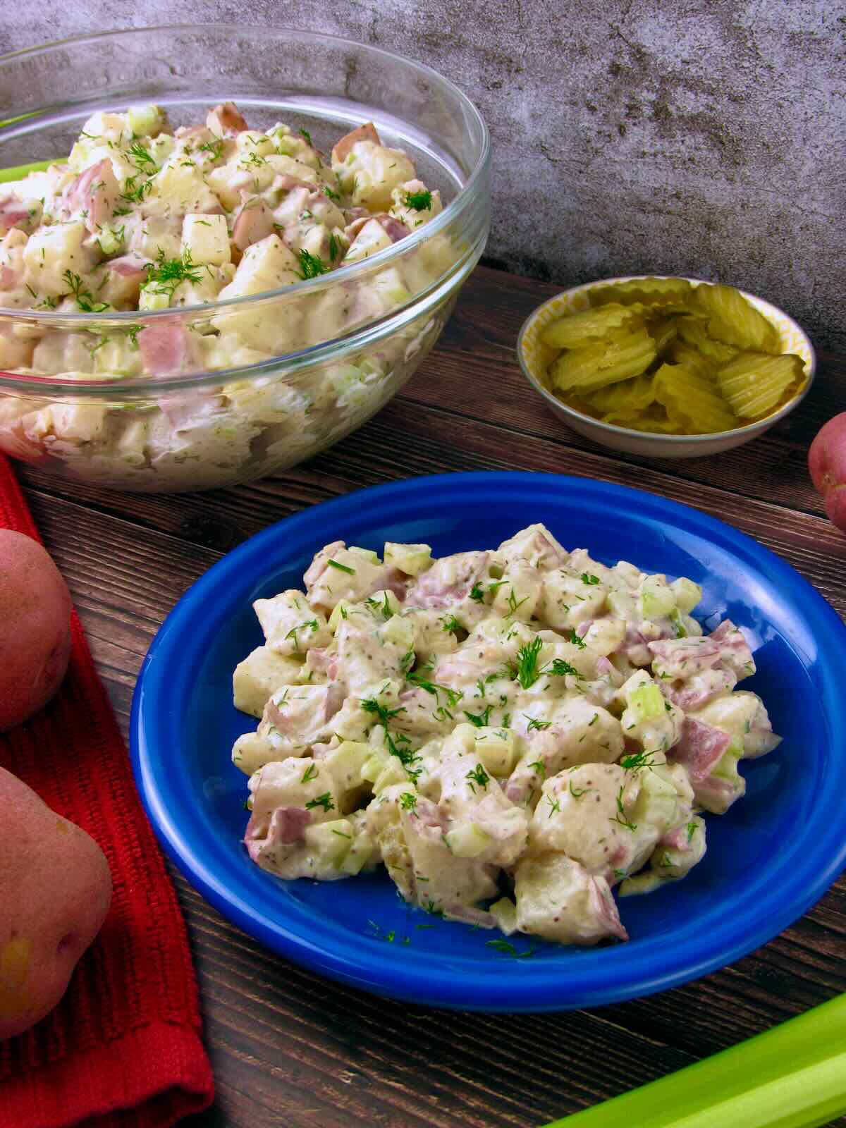 Dairy-free Vegan Potato Salad with dill served on a blue plate, large bowl and pickles in the background.