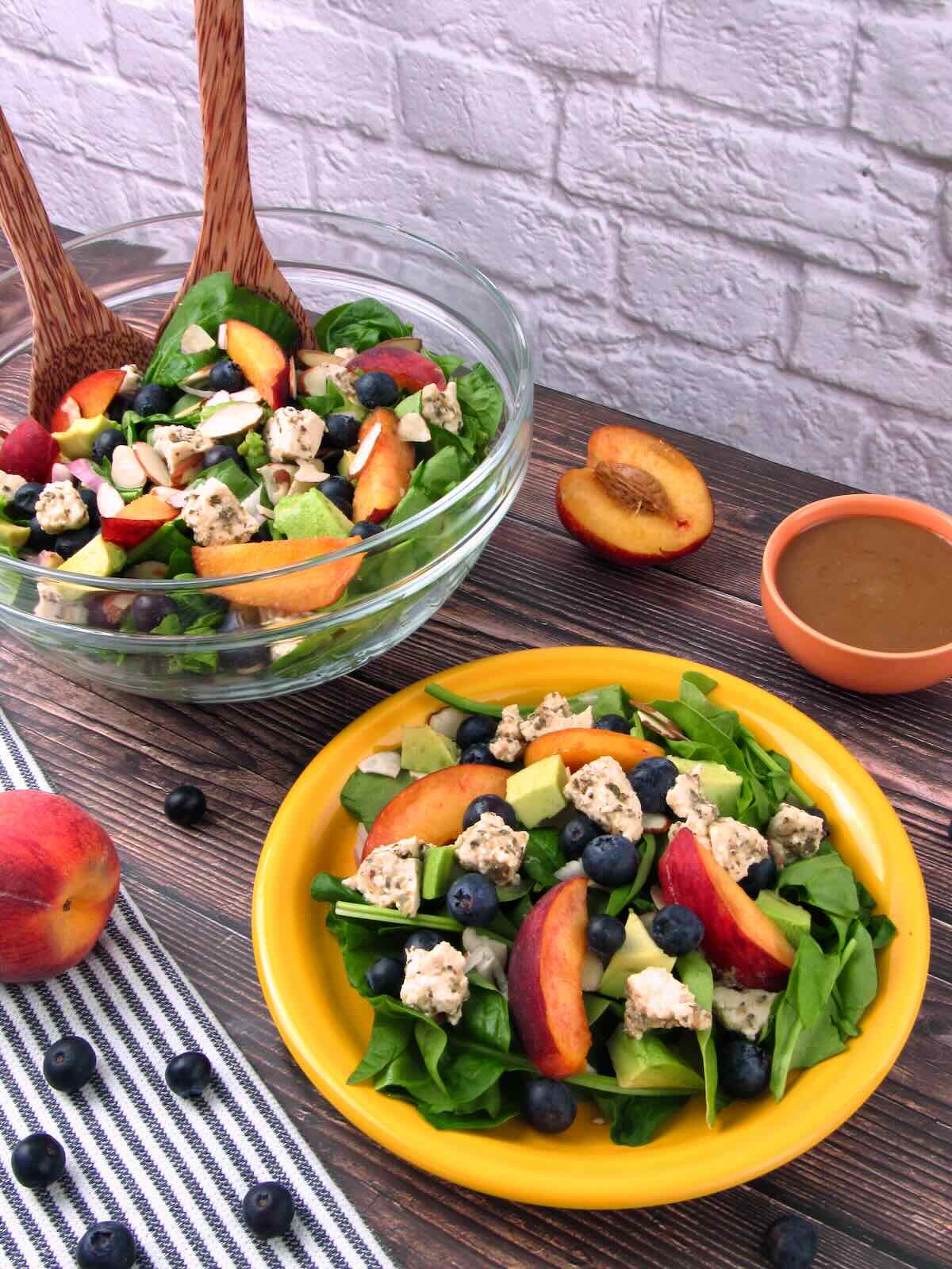 Peach Blueberry Spinach Salad served on a yellow plate, with a serving bowl of salad, fresh fruit, and balsamic dressing in the background.