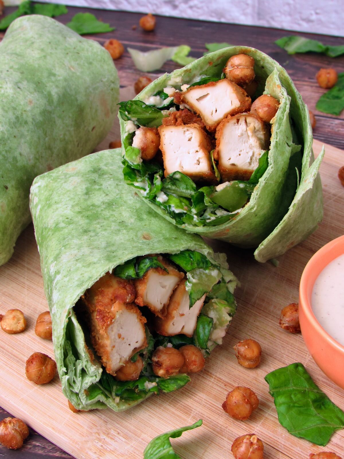 Spinach tortilla Caesar Wrap filled with crispy chickpeas, baked tofu chicken, chopped romaine, and creamy Caesar dressing, sliced in half.