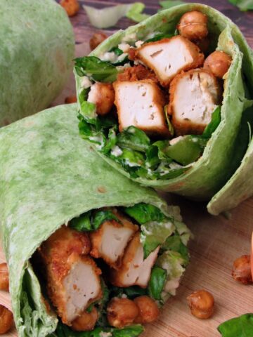 Close-up of a Vegan Chicken Caesar Wrap made with spinach tortilla, crispy tofu chicken, roasted chickpeas, chopped romaine, and creamy dressing, served on a wooden board with a small bowl of dressing on the side.