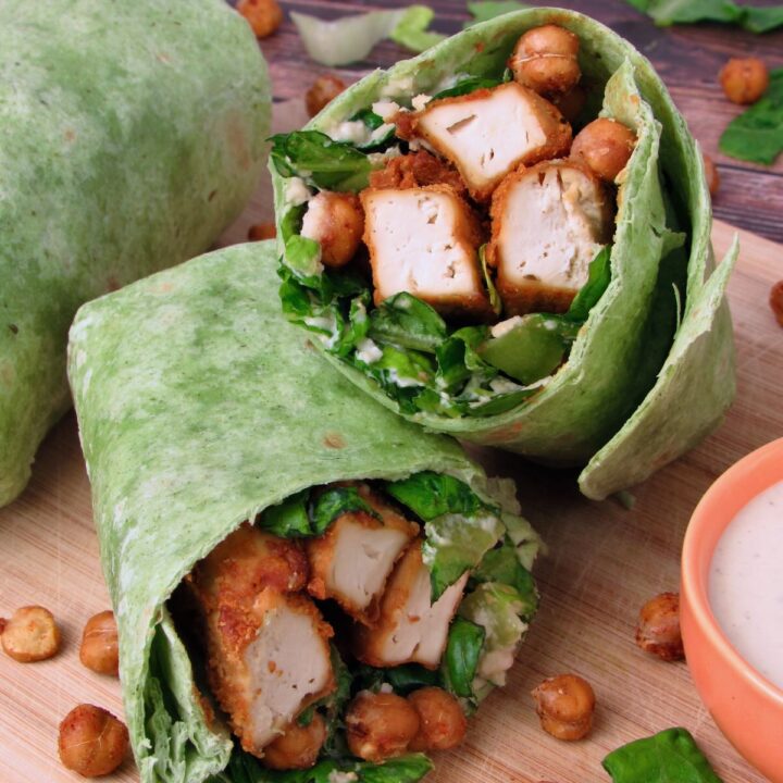 Close-up of a Vegan Chicken Caesar Wrap made with spinach tortilla, crispy tofu chicken, roasted chickpeas, chopped romaine, and creamy dressing, served on a wooden board with a small bowl of dressing on the side.