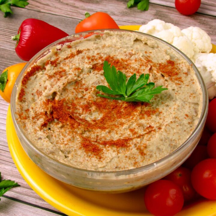 Bowl of oil-free Baba Ganoush topped with paprika and parsley, surrounded by cherry tomatoes, cauliflower, and colorful bell peppers on a yellow plate.