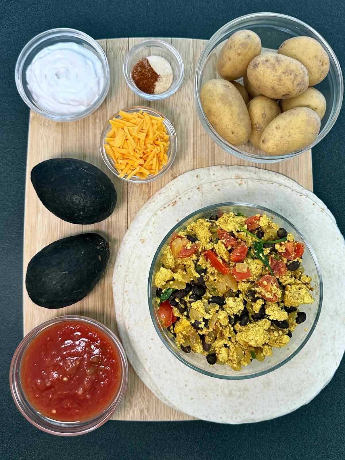 Ingredients for Tex Mex Vegan Breakfast Burritos arranged on wooden cutting board: bowl of tofu scramble, tortillas, potatoes, shredded vegan cheese, salsa, coconut yogurt, spices, and avocados.