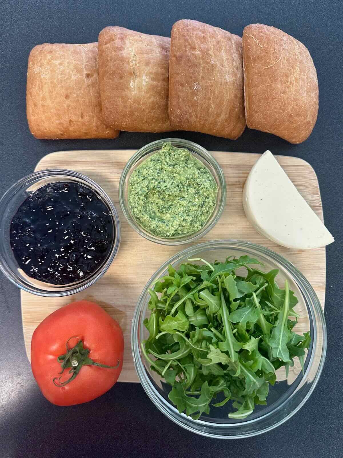 Vegan Pesto Sandwich ingredients including artisan bread, basil pesto, plant-based mozzarella, and fresh arugula on cutting board.