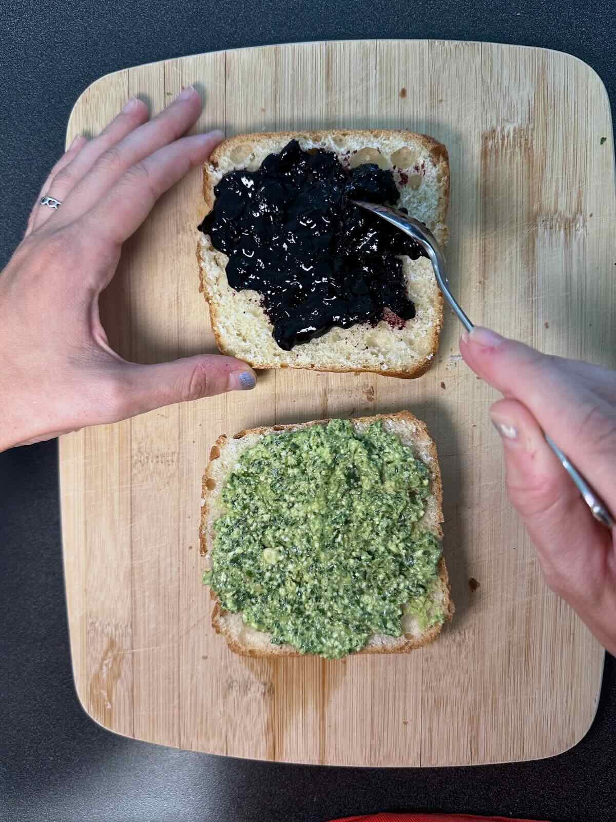 Spreading fresh basil pesto and balsamic blueberry jam on ciabatta bread for vegan sandwich preparation.