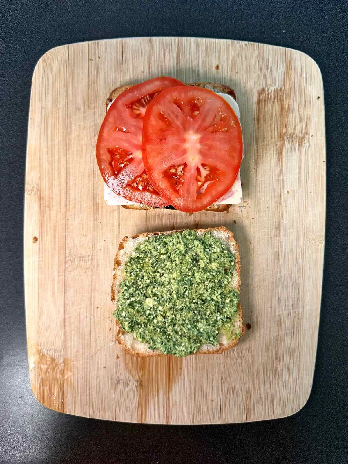 Open ciabatta bread with basil pesto on one half and blueberry jam, vegan mozzarella, and tomato slices on the other half.