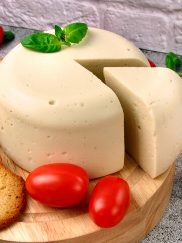 Easy Vegan Mozzarella Cheese on a wooden board with a wedge cut out, surrounded by toasted bread slices, cherry tomatoes, and fresh basil leaves.