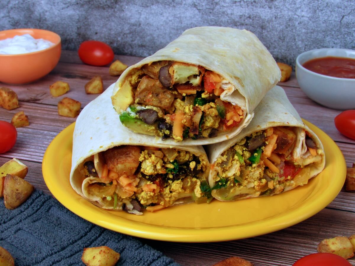 Three Tex Mex tofu scramble breakfast burritos stacked on a yellow plate with salsa and roasted potatoes.