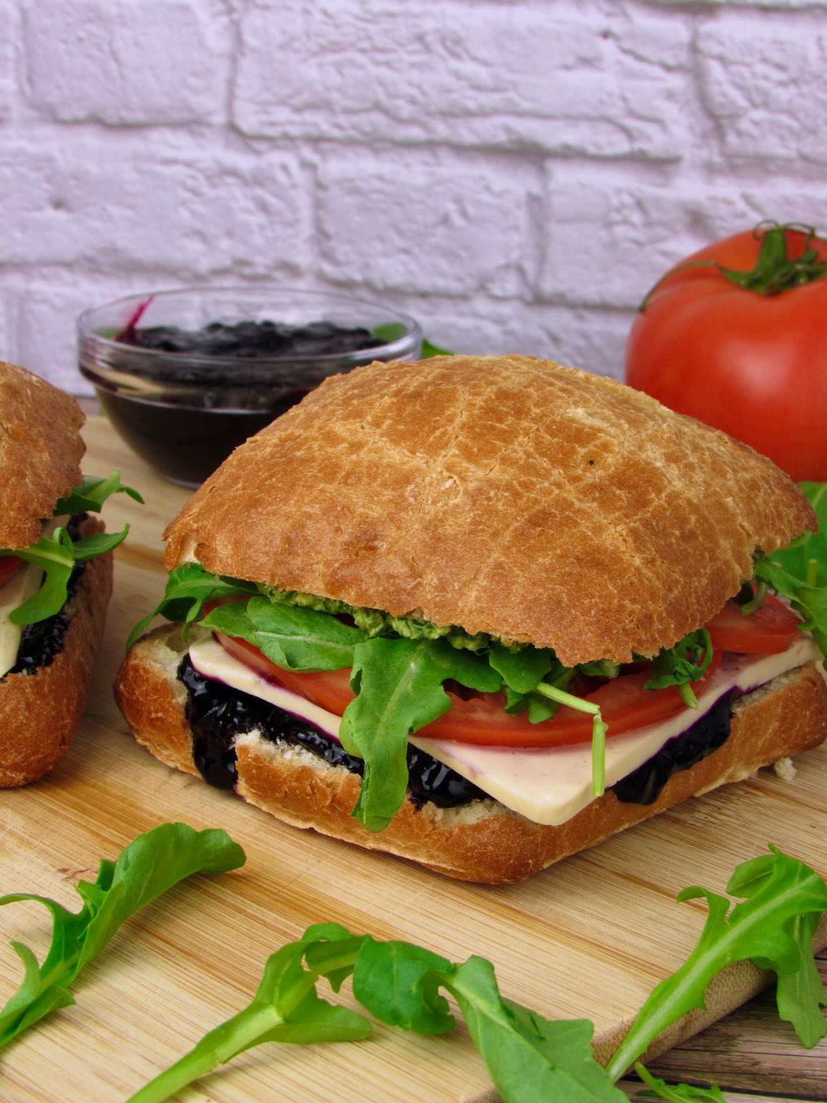 Vegan Mozzarella Pesto Sandwich with Blueberry Jam on crusty artisan bread with fresh arugula, tomato slices, and dark blueberry spread on a wooden cutting board.