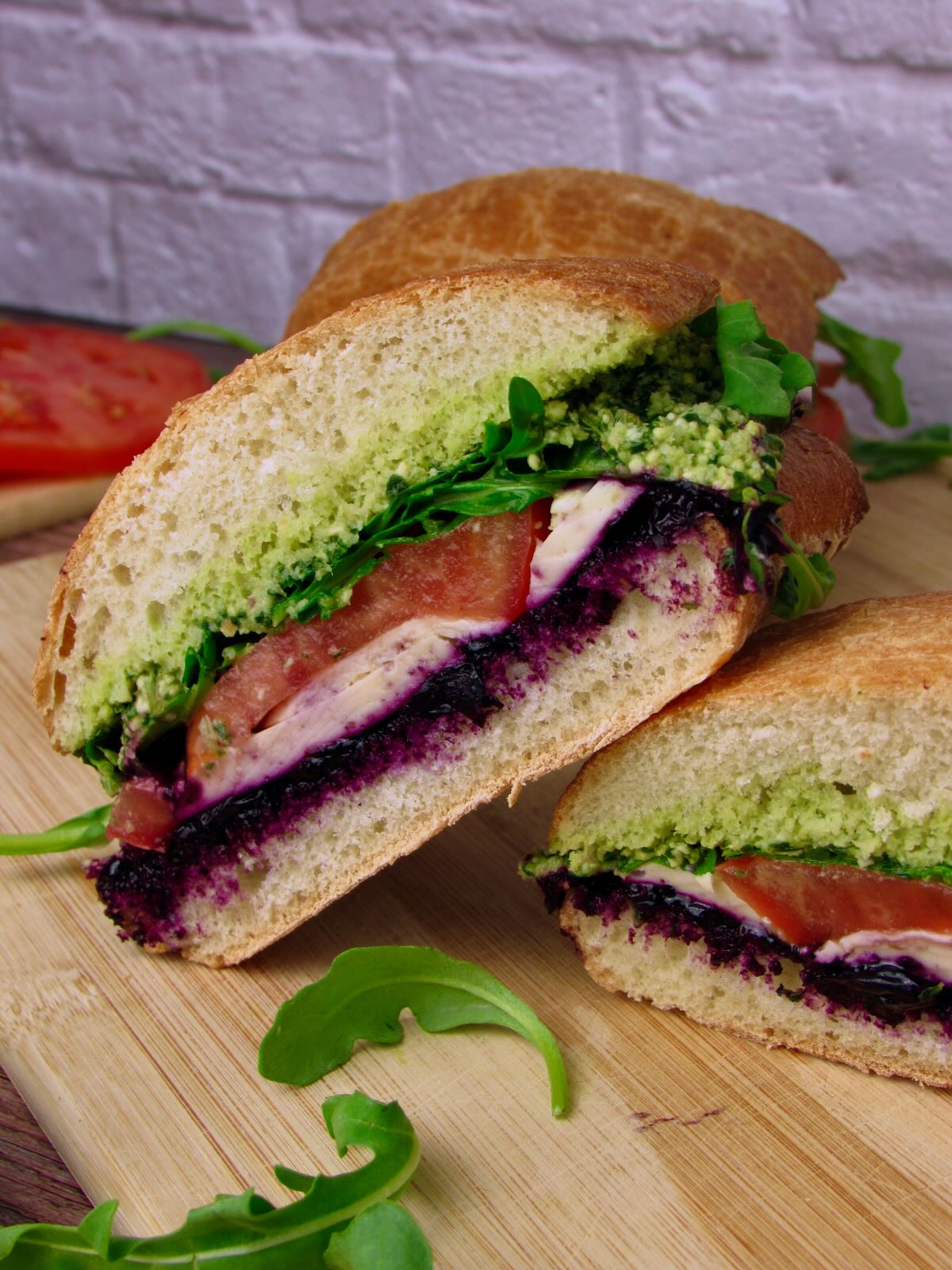 A Vegan Mozzarella Pesto Sandwich with Blueberry Jam showing layers of basil pesto, vegan mozzarella, balsamic blueberry jam, tomato, and arugula on ciabatta bread.