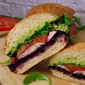 A halved Vegan Mozzarella Pesto Sandwich with Blueberry Jam showing colorful layers of pesto, vegan mozzarella, blueberry jam, tomato, and arugula on crusty bread.