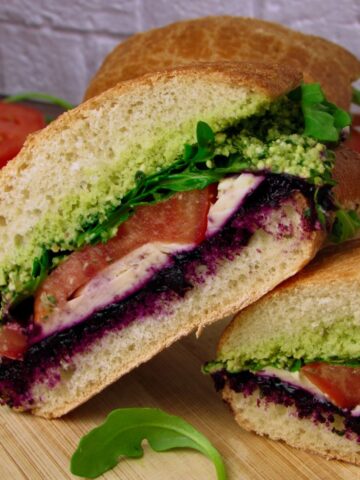 A halved Vegan Mozzarella Pesto Sandwich with Blueberry Jam showing colorful layers of pesto, vegan mozzarella, blueberry jam, tomato, and arugula on crusty bread.
