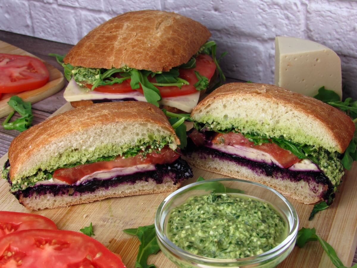 Vegan Sandwich with basil pesto, plant-based mozzarella, and blueberry jam cut in half on a wooden cutting board surrounded by basil pesto, ripe tomatoes, and fresh arugula.