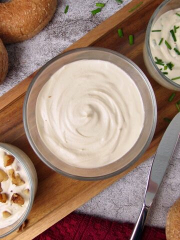 Three bowls of Vegan Cashew Cream Cheese, plain, chive, and maple walnut, on a wooden board with bagels and a butter knife.
