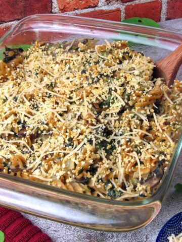 Vegan Mushroom Spinach Pasta Bake in a glass baking dish topped with golden breadcrumbs and shredded vegan cheese, with a wooden spoon scooping into the casserole.