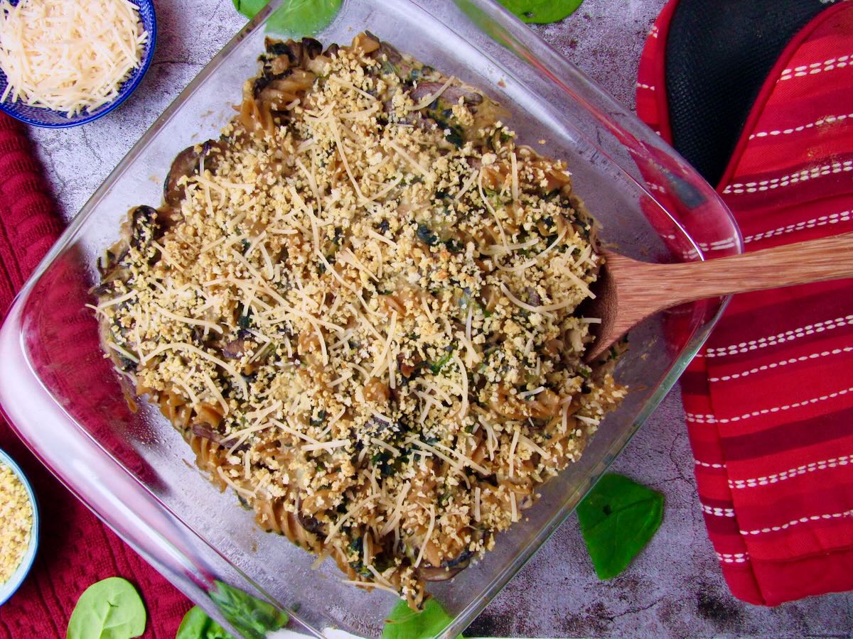 Overhead shot of Creamy Vegan Mushroom Spinach Pasta Bake in a casserole dish with a wooden spoon, a cozy plant-based comfort food dinner topped with breadcrumb topping.