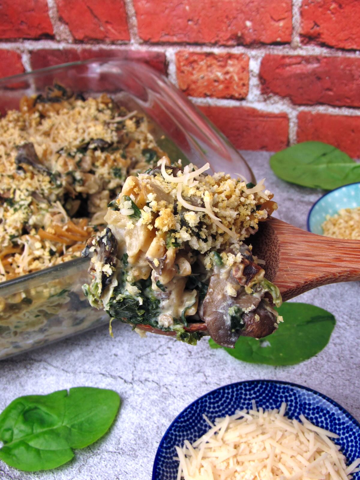 Creamy Vegan Mushroom Spinach Pasta Bake being scooped onto a wooden spoon, with spinach and vegan parmesan nearby.