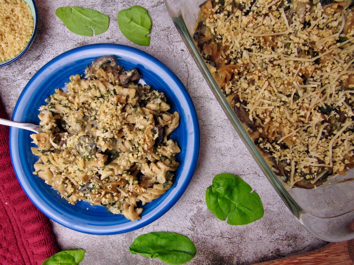Serving of Vegan Mushroom Spinach Pasta Bake on a blue plate topped with crispy breadcrumbs, with the casserole dish and fresh spinach leaves nearby.
