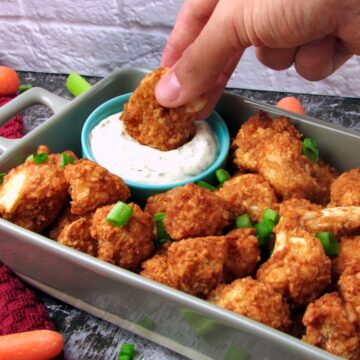 Hand dipping a crispy breaded Baked Buffalo Cauliflower Wing into Vegan Ranch Dressing, surrounded by more golden cauliflower bites and veggies on a platter.