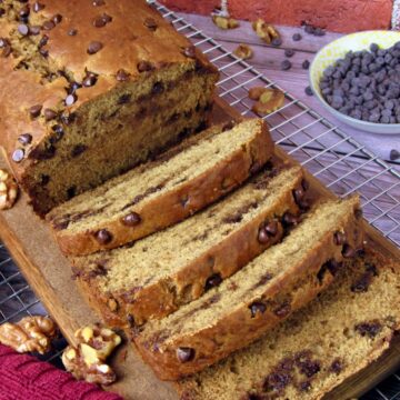 Sliced loaf of vegan banana bread with chocolate chips on a wooden board, cooling on a wire rack with walnuts and extra chocolate chips scattered around.
