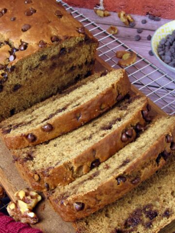 Sliced loaf of vegan banana bread with chocolate chips on a wooden board, cooling on a wire rack with walnuts and extra chocolate chips scattered around.