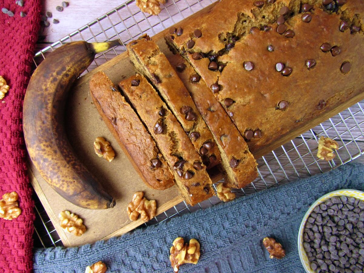 Overhead view of a banana bread loaf, partially sliced with a ripe banana and walnuts nearby.