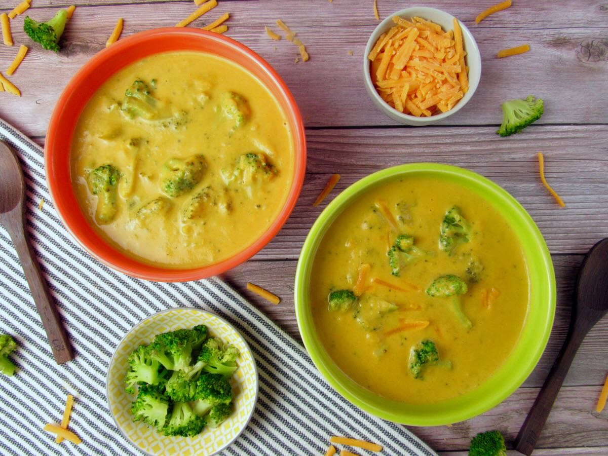 Overhead view of Dairy-Free Broccoli Cheese Soup, surrounded by a wooden spoon, vegan cheddar cheese, and steamed broccoli.