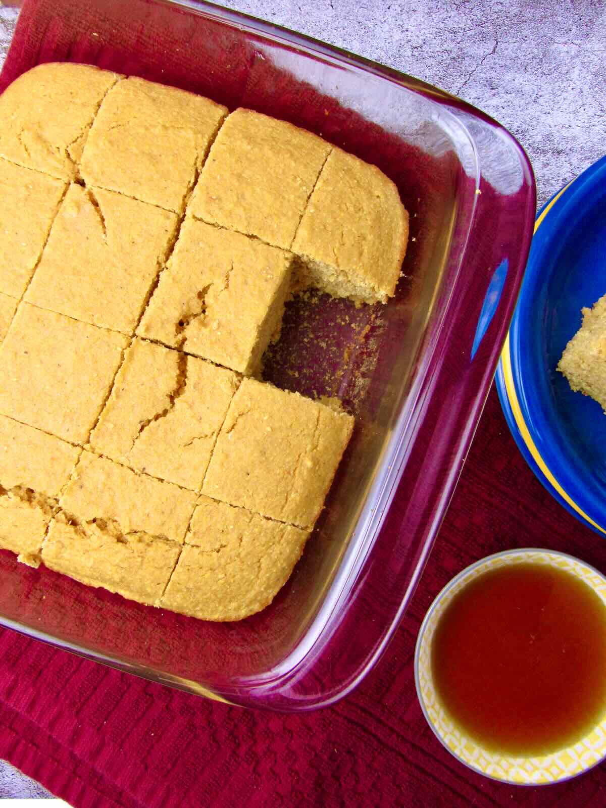 Sliced homemade cornbread with a piece missing, with maple syrup served on the side.