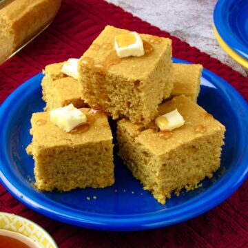 Moist vegan cornbread squares stacked on a blue plate, topped with vegan butter and maple syrup.