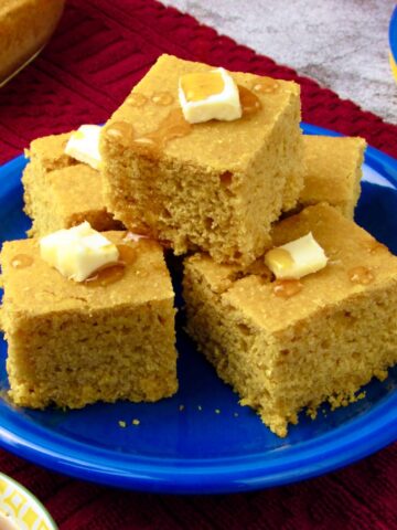 Moist vegan cornbread squares stacked on a blue plate, topped with vegan butter and maple syrup.