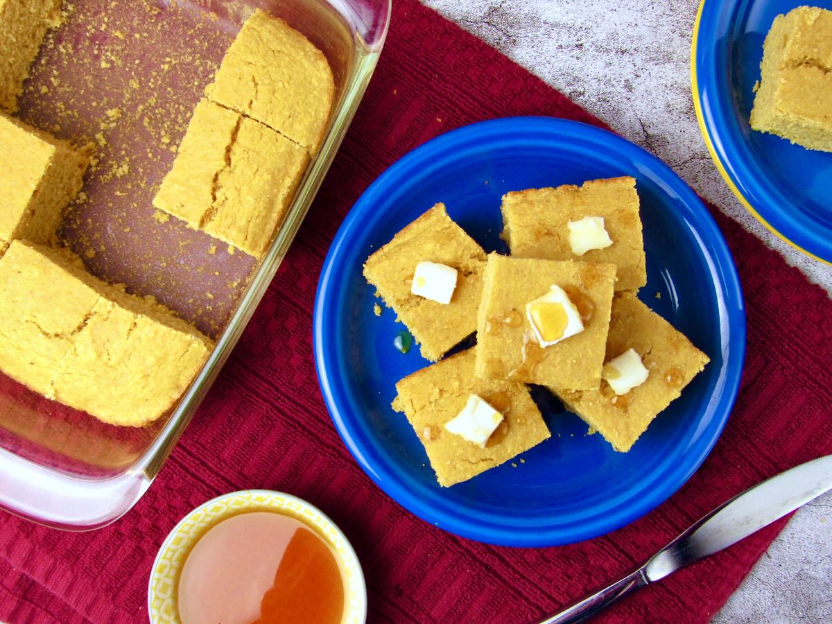 Overhead view of cornbread cut into squares, with several pieces on a blue plate topped with vegan butter and syrup, next to a baking dish of remaining cornbread.