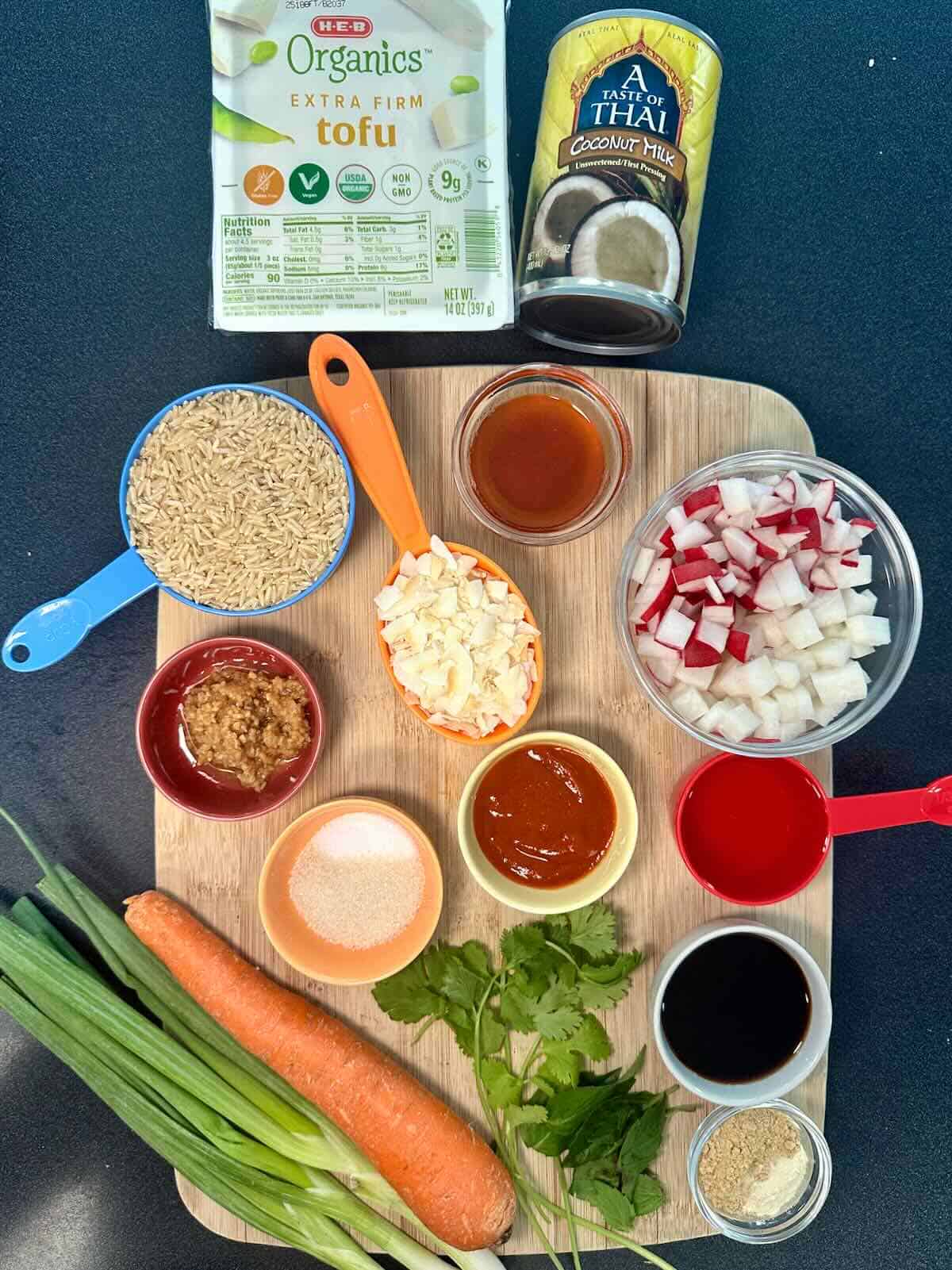 Overhead view of ingredients for a coconut rice bowl including tofu, jasmine rice, pickled radish, carrots, coconut milk, fresh herbs, and seasonings.