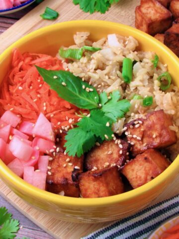 Zesty Coconut Rice Buddha Bowl with crispy sriracha tofu, quick pickled radish, shredded carrots, sesame seeds, and fresh herbs in a yellow bowl.