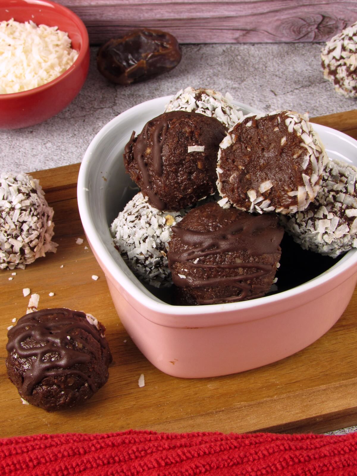 Chocolate coconut date truffle bites with a dark chocolate drizzle and coconut coating, served in a small pink bowl on a wooden board.