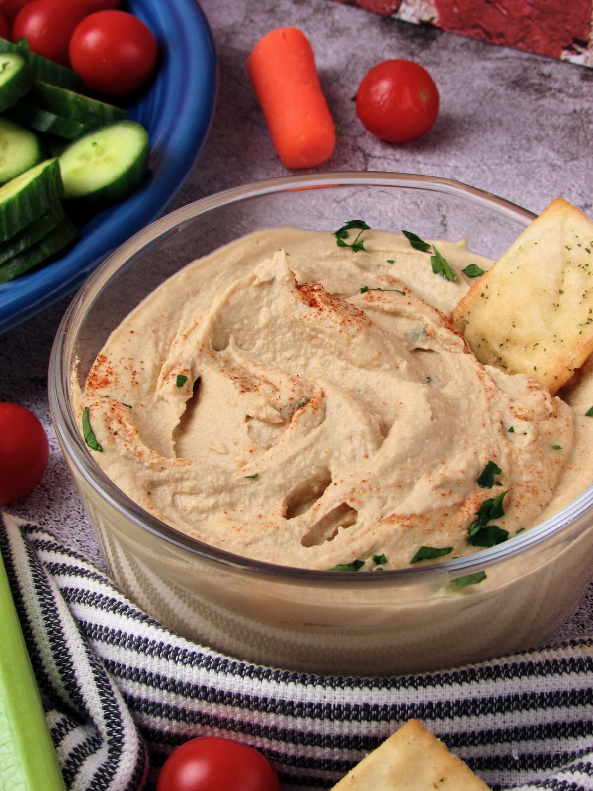 Bowl of smooth garlic hummus garnished with paprika and fresh parsley, served with a pita cracker and fresh veggies.