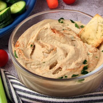 Creamy homemade garlic hummus in a bowl with a pita cracker dipped in, garnished with paprika and fresh parsley.