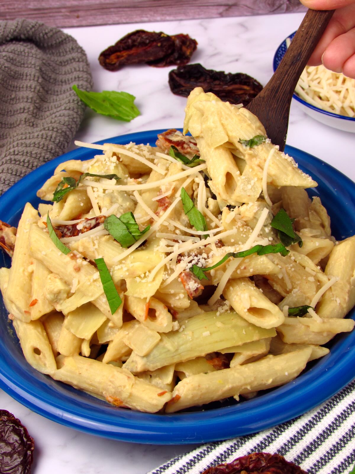 Side view of creamy lemon hummus pasta with artichokes and sun-dried tomatoes, with a fork lifting a bite.