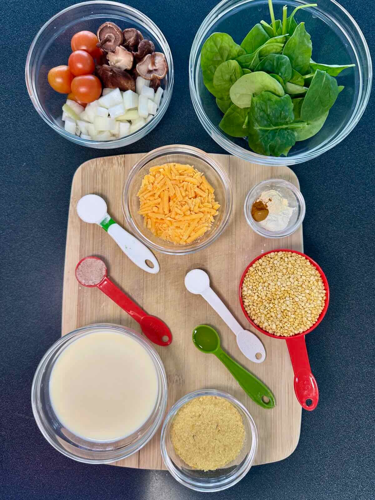 Ingredients for a vegan mung bean omelet, including mung beans, plant milk, nutritional yeast, vegan shredded cheese, cherry tomatoes, mushrooms, diced onion, and fresh spinach.