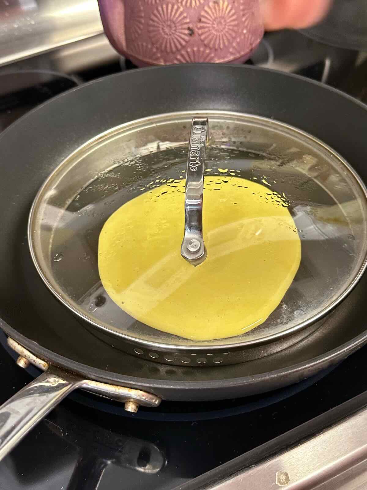 Vegan mung bean omelet batter cooking in a skillet with a lid on.