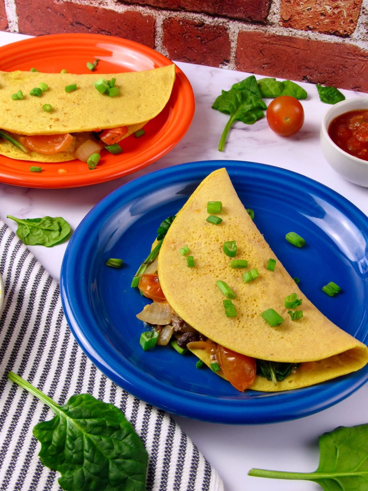 Vegan mung bean omelet filled with sautéed mushrooms, tomatoes, onion, and spinach, topped with green onions and served on colorful plates.