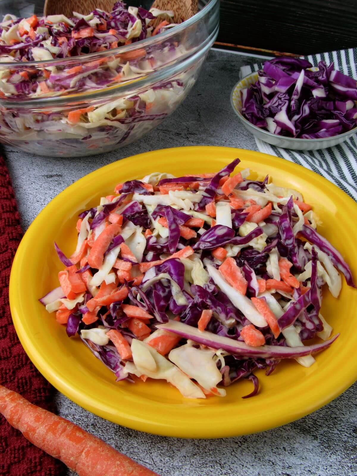 Creamy vegan coleslaw served on a yellow plate, with cabbage and carrots around it.