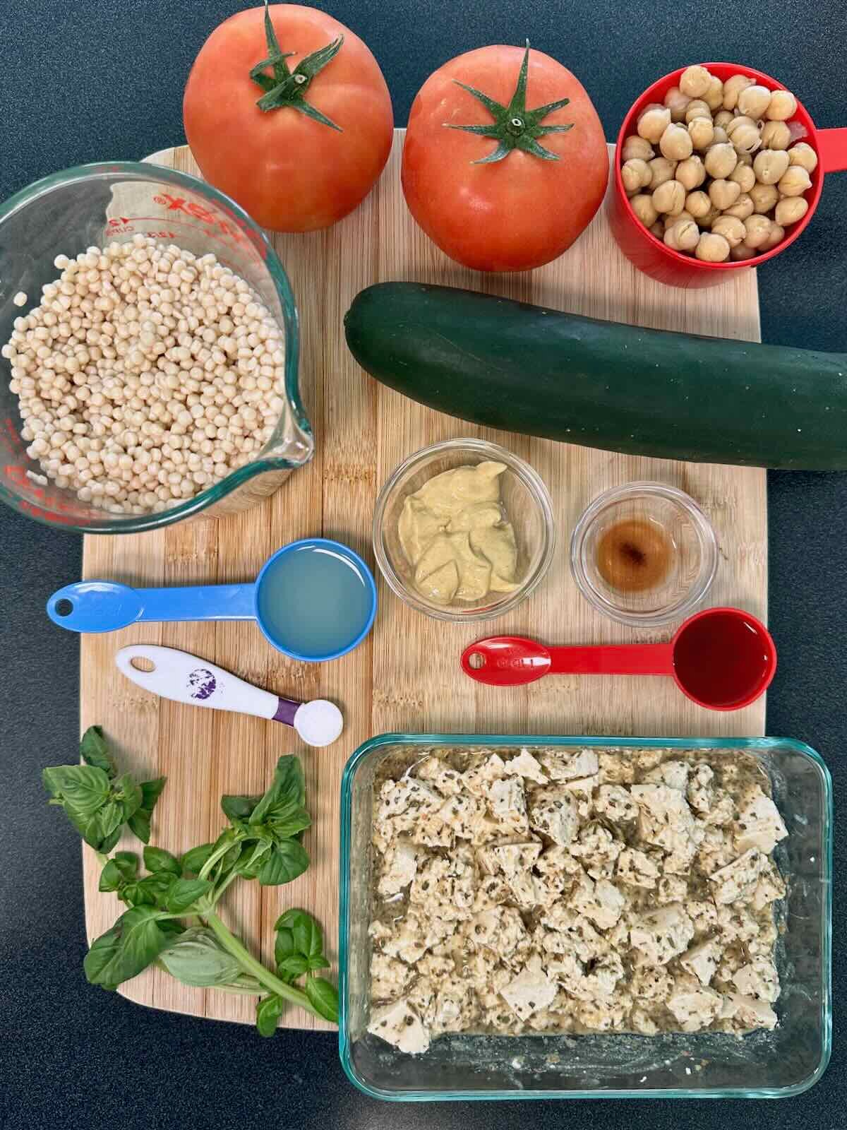 Ingredients for a Summer Couscous Salad, including cucumber, tomatoes, marinated vegan feta, fresh basil, and cooked pearl couscous.