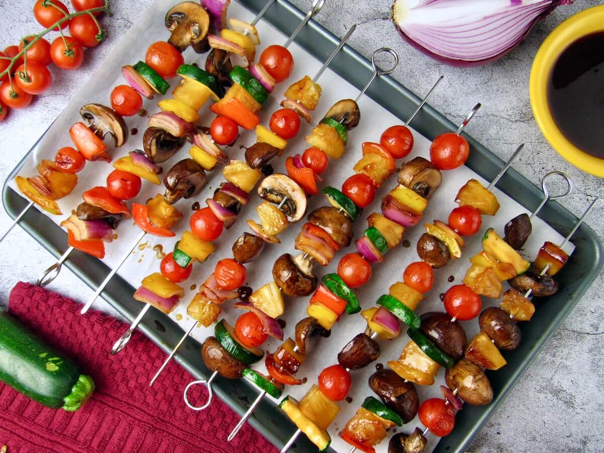 Overhead of fresh vegetable skewers on a baking tray, surrounded by cherry tomatoes, zucchini, and red onion.