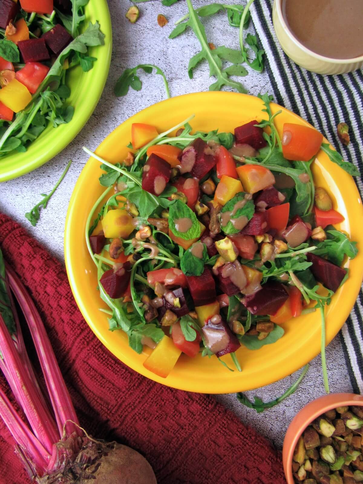 Overhead view of a roasted beet and arugula salad on a plate, topped with juicy tomatoes, chopped pistachios, fresh mint, and drizzled with citrus tahini dressing.